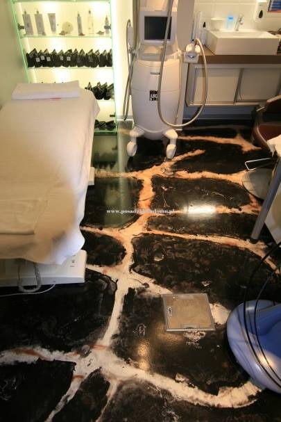 Nowoczesny gabinet kosmetyczny z marmurową posadzką żywiczną, białym łóżkiem zabiegowym i specjalistycznym sprzętem. Widoczna przeszklona szafka z kosmetykami.