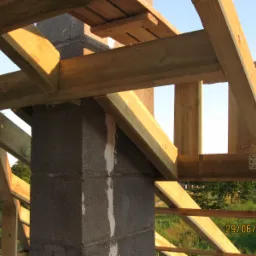 Wiązanie drewnianych belek dachowych z kominem z bloczków betonowych. Widoczne metalowe łączniki i fragment krajobrazu w tle. Konstrukcja w surowym stanie.