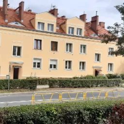 Odnowiony budynek wielorodzinny z żółtą elewacją, czerwoną dachówką i białymi obramowaniami okien, widziany zza żywopłotu i żółtej bariery drogowej.