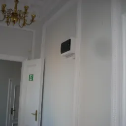 Białe wnętrze po pracach, z widocznym urządzeniem na ścianie, prawdopodobnie po usunięciu wilgoci. Widoczne drzwi z zielonym znakiem ewakuacyjnym i ozdobny żyrandol.