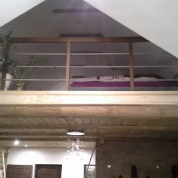 Wnętrze domu z bali z antresolą, widok z dołu na drewniany strop i balustradę z metalowymi prętami, na antresoli widoczne łóżko i rośliny doniczkowe.