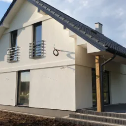 Nowoczesny dom jednorodzinny z jasną elewacją, ciemną dachówką i drewnianymi słupami podtrzymującymi zadaszenie wejścia, na etapie wykończenia terenu wokół budynku.