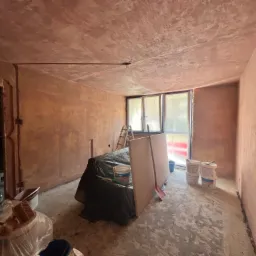 Pokój w trakcie przygotowania do prac wykończeniowych: ściany z nałożoną warstwą gładzi, zabezpieczone meble folią, widoczne wiadra z masą szpachlową i drabina oparta o okno.