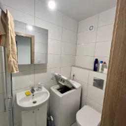 Nowoczesna, jasna łazienka z białą armaturą: umywalka z szafką, pralka z otwartym bębnem, wisząca toaleta i lustro z drewnianą ramą na tle białych płytek.