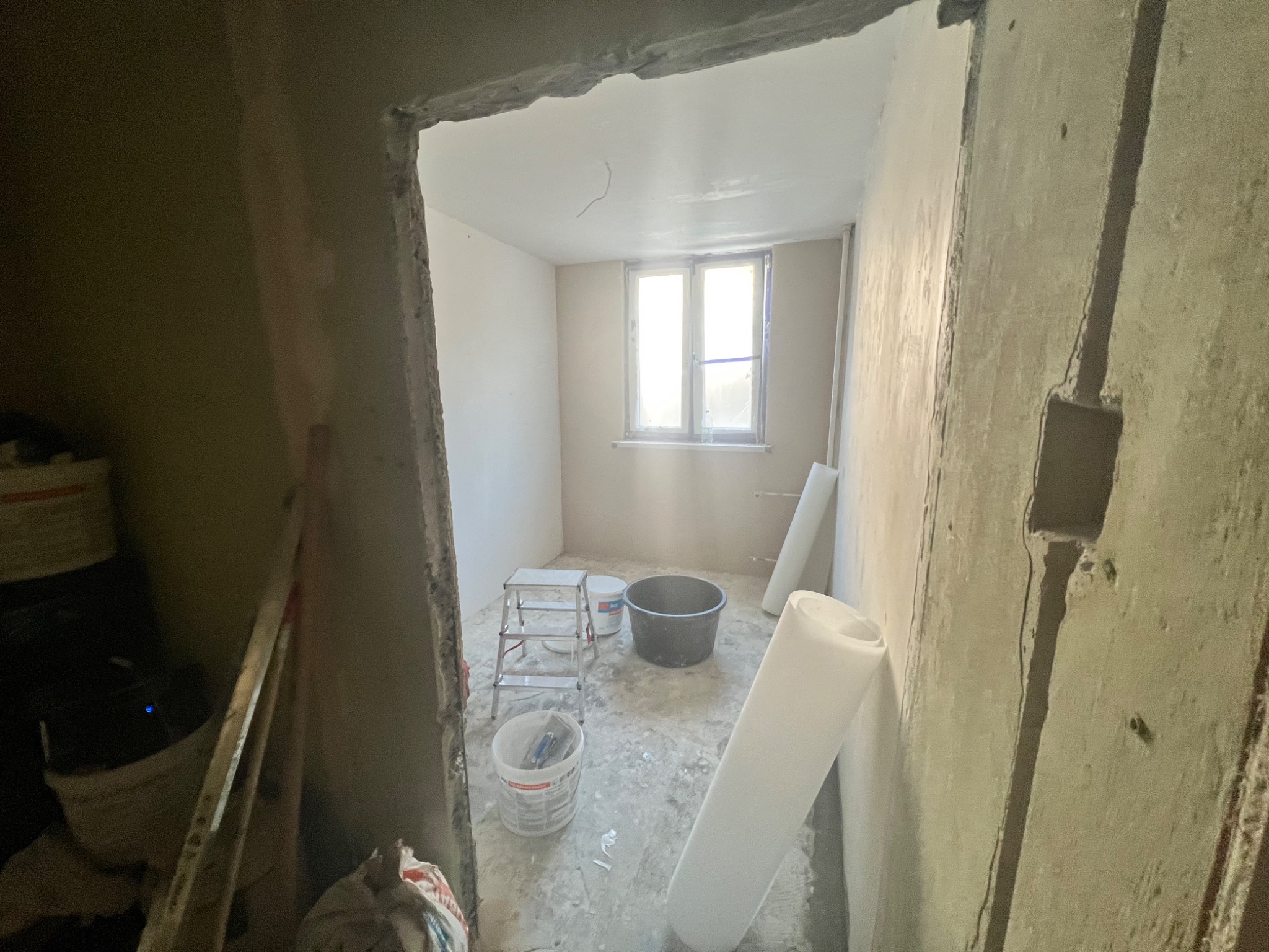 Wnętrze pokoju w Jeleśni w trakcie prac przygotowawczych do wykończenia ścian, widoczne surowe ściany, drabina, wiadro z narzędziami i rolka materiału.