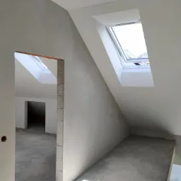 Poddasze w trakcie wykańczania: widoczne surowe ściany z otworami okiennymi, brak wykończenia podłogi, przygotowane otwory drzwiowe z widoczną instalacją elektryczną.