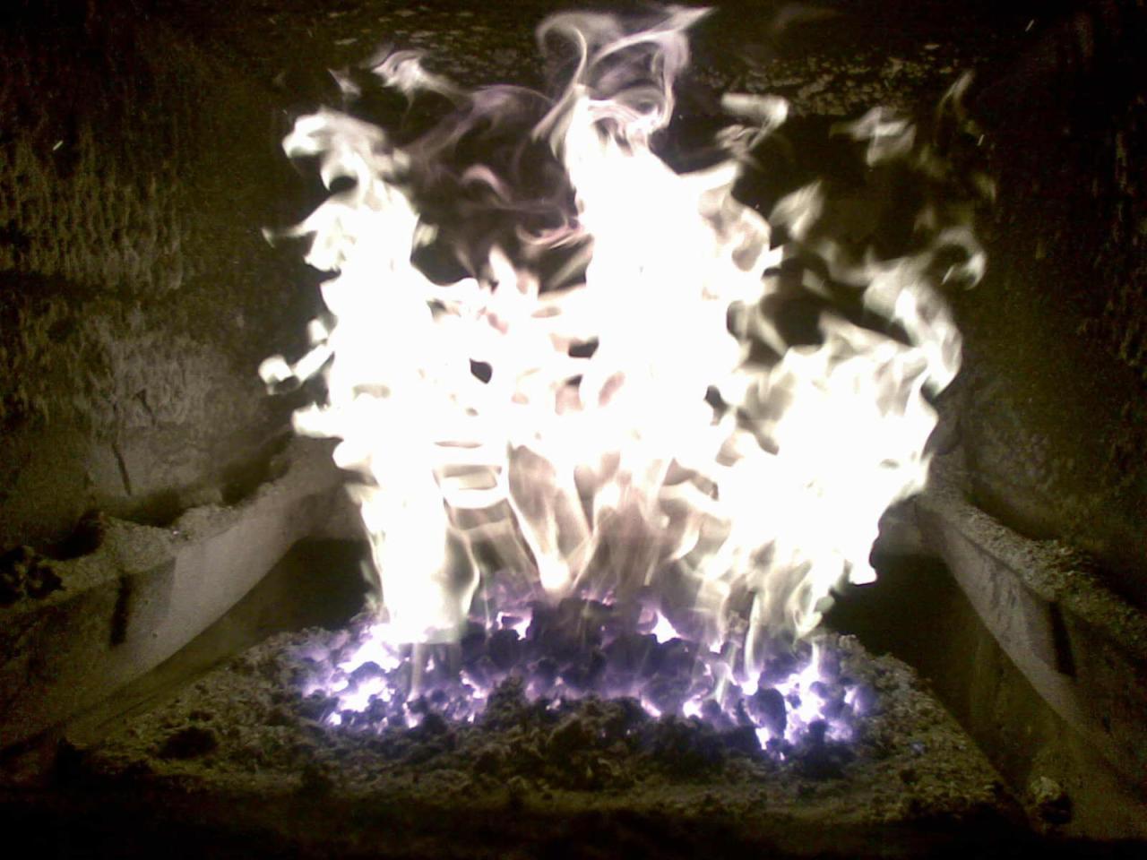 Płonące paliwo stałe w piecu, widoczne intensywne płomienie i żarzące się fragmenty, wnętrze pieca o ciemnej, szorstkiej powierzchni.