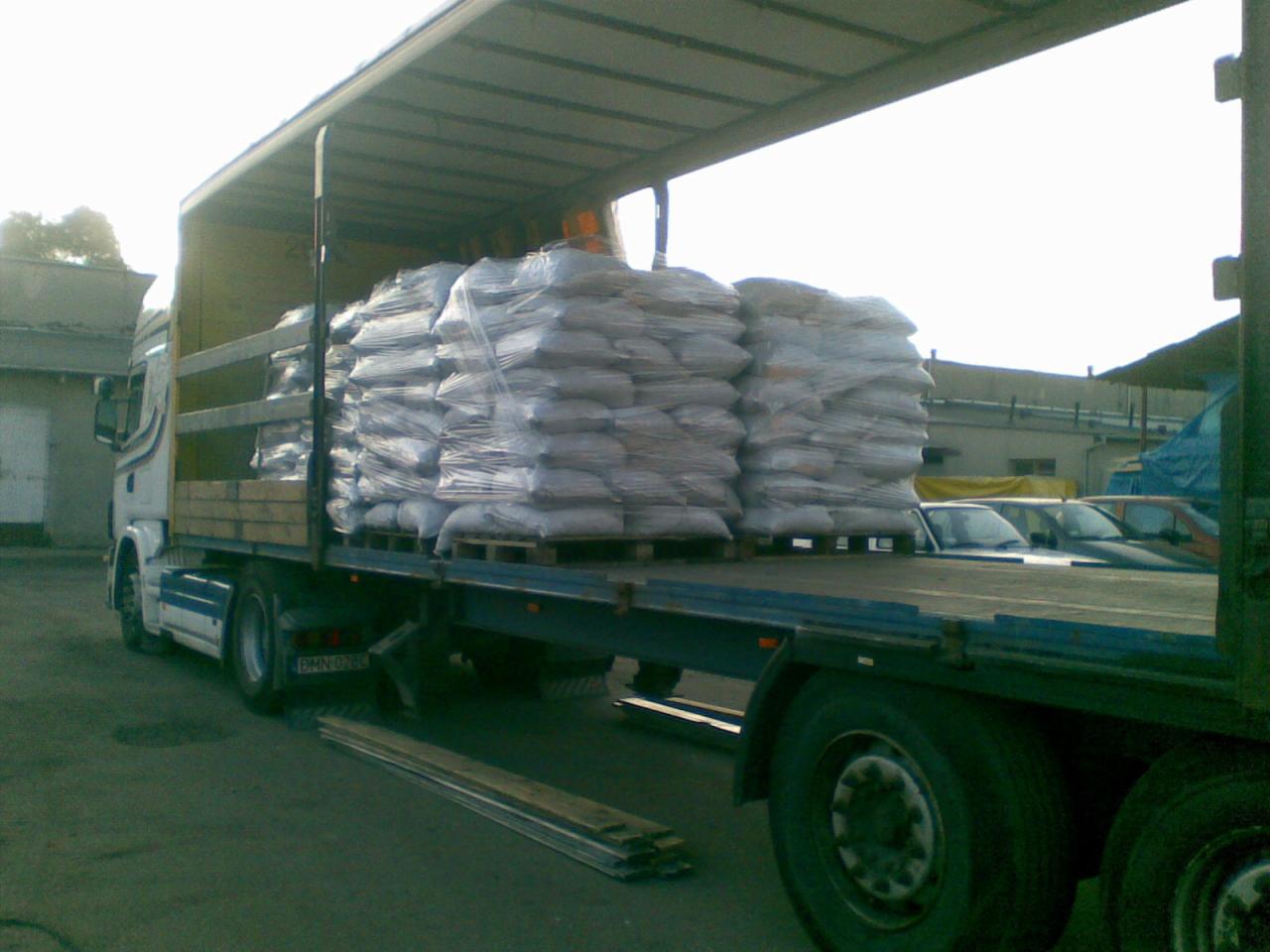 Ciężarówka z naczepą załadowana paletami z workowanym materiałem opałowym, zabezpieczonym folią stretch, w trakcie rozładunku na placu.