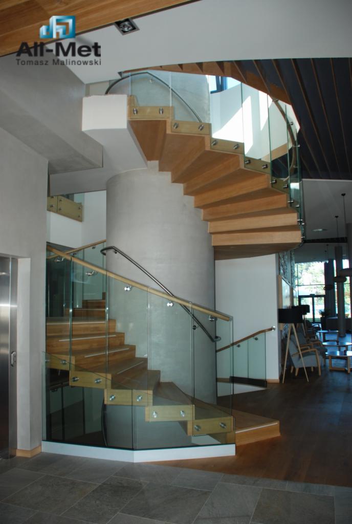 Spiralne schody z drewnianymi stopniami i szklaną balustradą, wsparte na centralnym betonowym filarze, oświetlone naturalnym światłem w nowoczesnym wnętrzu.