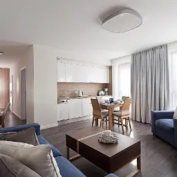 Jasne wnętrze apartamentu z błękitną sofą, drewnianym stołem jadalnym i zasłonami w odcieniach szarości wpadających do mieszkania przez okno.