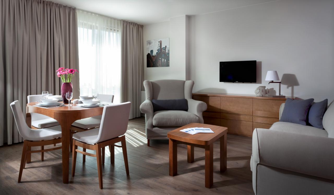 Jasne wnętrze apartamentu z zasłoniętymi oknami, drewnianym stołem jadalnym z nakryciem, wygodnym fotelem i sofą oraz telewizorem na ścianie.