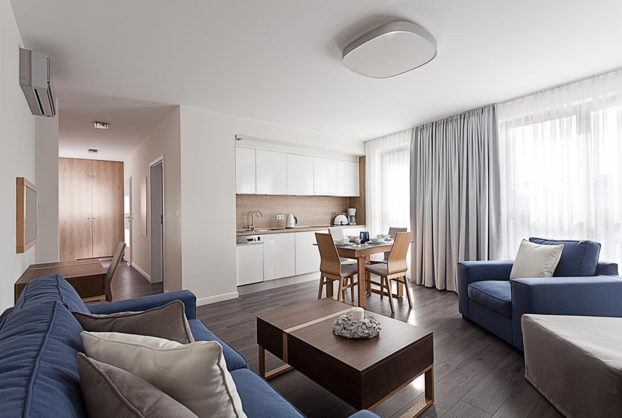 Jasne wnętrze apartamentu z błękitną sofą, drewnianym stołem jadalnym i zasłonami w odcieniach szarości wpadających do mieszkania przez okno.