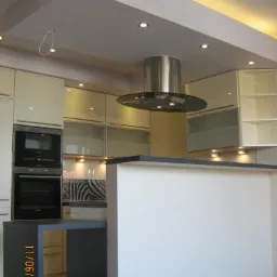 Nowoczesna kuchnia w jasnych barwach z wyspą kuchenną, oświetleniem punktowym i okapem cylindrycznym.