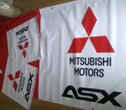 Biały baner reklamowy Mitsubishi Motors ASX z czerwonym logo i napisem, leżący na czerwonym materiale, obok mniejsze, podobne banery.