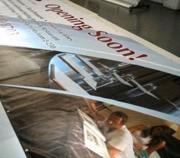 Mężczyzna montuje duży baner reklamowy z napisem 'Opening Soon!' i grafiką budynku. Widoczne fragmenty innych banerów w tle.