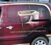 Boczny widok samochodu oklejonego folią z reklamą farb lateksowych i efektów dekoracyjnych. Na folii widoczny jest pojemnik z farbą marki Magnat w odcieniach brązu i beżu.