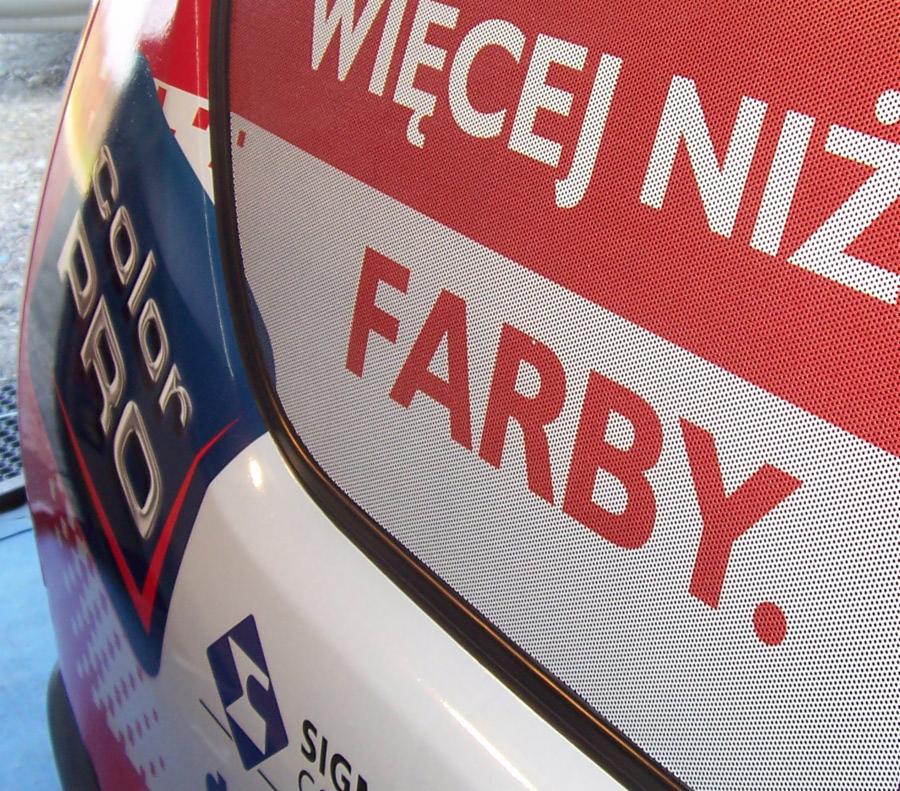 Fragment oklejonego samochodu z perforowaną folią OWV z reklamowym napisem 'WIĘCEJ NIŻ FARBY' w kolorze czerwonym i logo Color PRO na boku pojazdu.