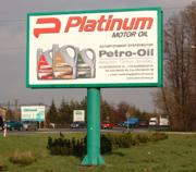 Billboard reklamowy z grafiką prezentującą oleje silnikowe marki Platinum Motor Oil i Petro-Oil, umieszczony na zielonym, metalowym słupie na tle miejskiego krajobrazu z przejeżdżającymi samochodami.