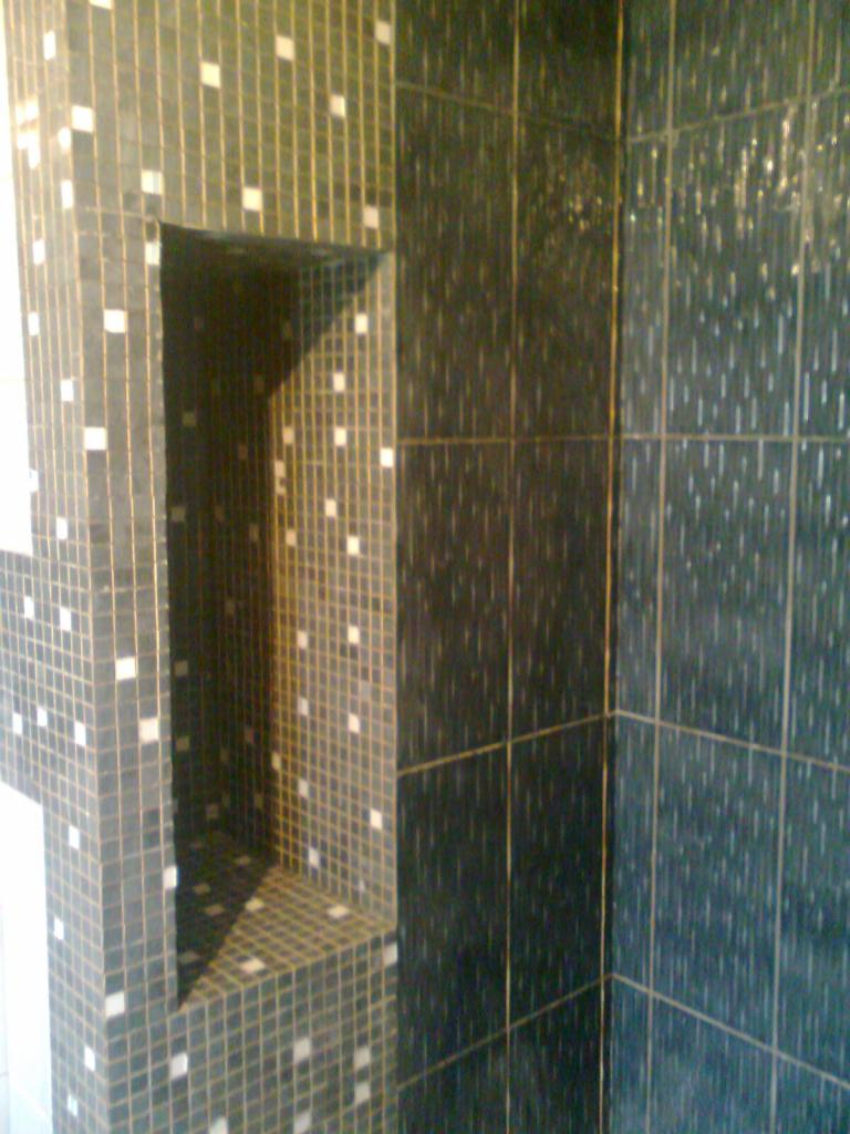 Fragment ściany łazienki wyłożonej ciemną mozaiką z jasnymi akcentami, wbudowana półka w ścianie, widoczne fugi.