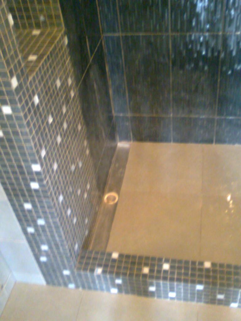 Wykończony brodzik prysznicowy z mozaiką w tonacji szarości i bieli oraz ciemnymi płytkami na ścianach, widoczny odpływ liniowy.