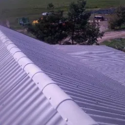 Szary dach z blachy trapezowej z półokrągłymi elementami wykończeniowymi, widok z góry, w tle zieleń i budynek.