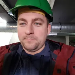 Portret inżyniera w zielonym kasku na tle rur wentylacyjnych pod sufitem.