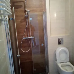 Nowoczesna łazienka z kabiną prysznicową z deszczownicą, toaletą wiszącą i grzejnikiem drabinkowym, wykończona płytkami imitującymi drewno i jasnym beżem.