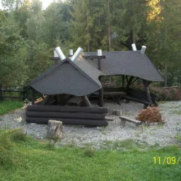 Drewniana altana ogrodowa z dwuspadowym dachem pokrytym ciemną dachówką bitumiczną na tle leśnym, z widocznymi detalami konstrukcyjnymi i elementami dekoracyjnymi.