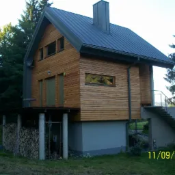 Nowoczesny dom o konstrukcji szkieletowej obłożony drewnem, z ciemnym dachem i kominem, posadowiony na betonowej podstawie, z zadaszonym tarasem i schodami wejściowymi, w otoczeniu drzew.