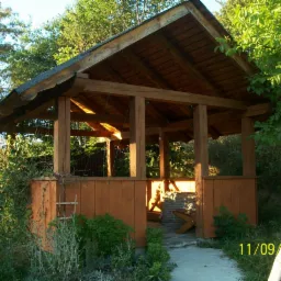 Drewniana altana z ciemnym dachem w ogrodzie, z widocznymi elementami konstrukcyjnymi, częściowo osłonięta drewnianą boazerią, z kamiennym grillem wewnątrz i ścieżką prowadzącą do wejścia.