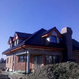 Niedokończony dom murowany z ciemną dachówką, widocznym kominem z cegły i stertą ziemi przed budynkiem, w słoneczny dzień.