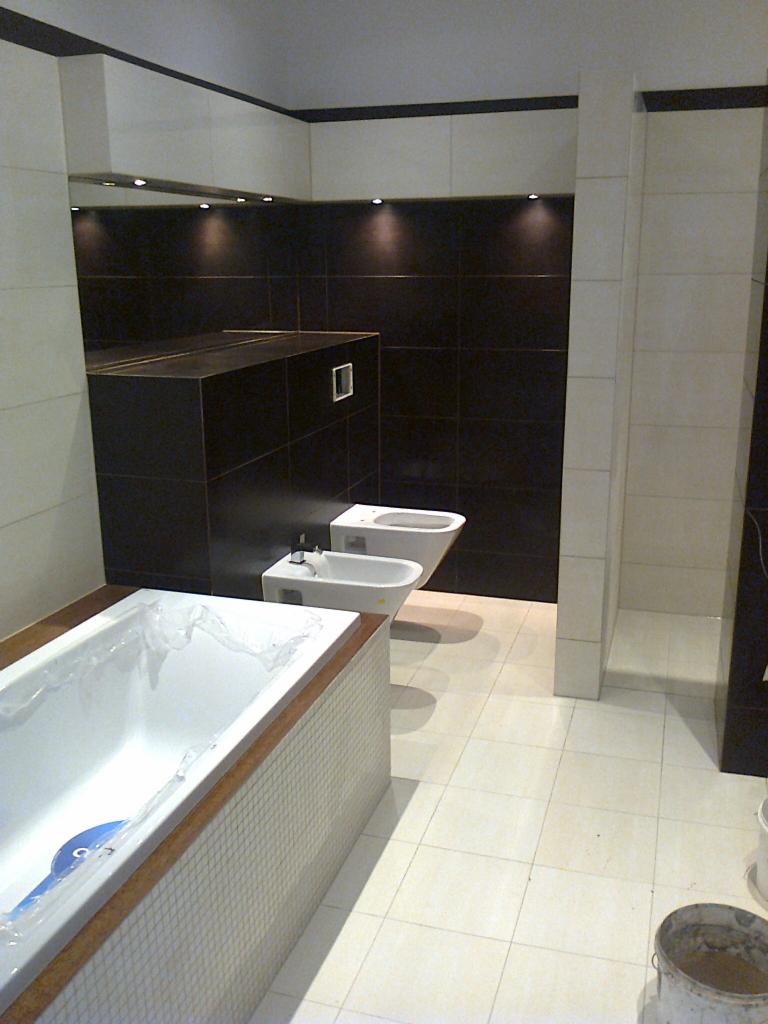 Nowoczesna łazienka z czarnymi płytkami na ścianach, białą wanną w folii ochronnej z mozaiką na obudowie, białym bidetem i toaletą oraz jasnymi płytkami podłogowymi.