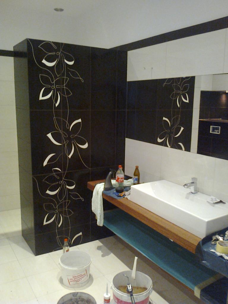 Czarno-biała łazienka z motywem kwiatowym na ciemnych płytkach, biała umywalka na drewnianym blacie, widoczne narzędzia i materiały budowlane w wiadrach na podłodze, sugerujące trwający proces...