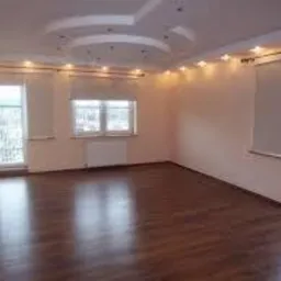 Pusty, jasny pokój z parkietem, oświetleniem punktowym w suficie podwieszanym i dwoma oknami balkonowymi z roletami rzymskimi.