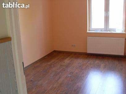 Wnętrze pokoju z drewnianą podłogą, grzejnikiem pod oknem i pomarańczowymi ścianami, widok z perspektywy wejścia.