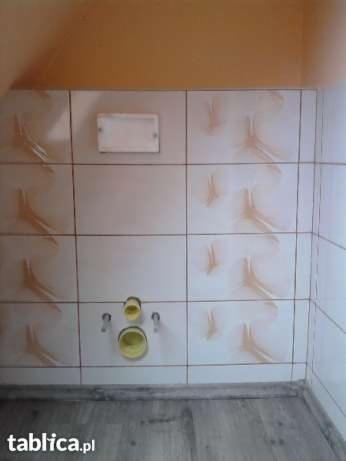 Ściana łazienki wyłożona kwadratowymi, beżowymi płytkami ceramicznymi z abstrakcyjnym, brązowym wzorem; widoczne otwory na instalację sanitarną i biała klapka rewizyjna.