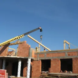Budowa domu z czerwonej cegły Porotherm. Żuraw podnosi drewniany element konstrukcyjny. Widoczny pracownik na górze budynku. W tle błękitne niebo.