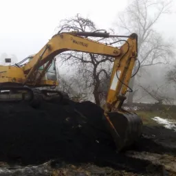 Żółta koparka Warynski na hałdzie czarnej ziemi w mglisty dzień, widoczne drzewa bez liści w tle.