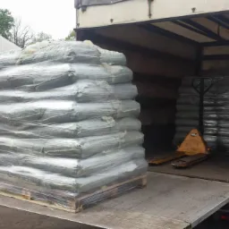 Załadunek workowanego ekogroszku na paletach do ciężarówki z windą. W tle widoczne zapasy i budynek magazynowy. Transport ekogroszku.