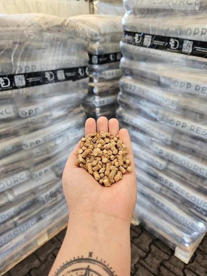 Garść pelletu drzewnego na tle zapakowanych worków z pelletem na paletach. Widoczny tatuaż kompasu na nadgarstku.