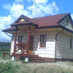 Niewielki, jasny dom z ciemnoczerwonym dachem bitumicznym i drewnianą werandą na betonowych podporach, widok z boku na tle błękitnego nieba z chmurami.