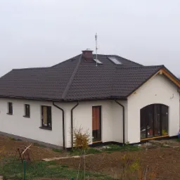 Gotowy, parterowy dom jednorodzinny z brązowym dachem z dachówki, białymi ścianami, oknami w ciemnych ramach i anteną na kominie, otoczony świeżo przygotowanym terenem pod ogród.