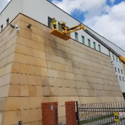 Pracownik w żółtym kombinezonie na podnośniku koszowym czyści elewację budynku pokrytą płytami z piaskowca. Widoczne ciemniejsze smugi na elewacji wskazują na obszar po czyszczeniu.