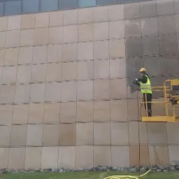 Pracownik w żółtym kasku i kamizelce na podnośniku koszowym czyści elewację budynku z jasnych, kwadratowych płyt, wyraźnie widoczna różnica w kolorze między czystą a brudną powierzchnią.