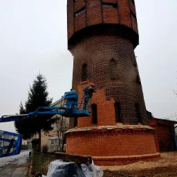 Renowacja ceglanej wieży ciśnień z użyciem podnośnika koszowego, widoczne ślady czyszczenia elewacji, częściowo odnowiona dolna partia