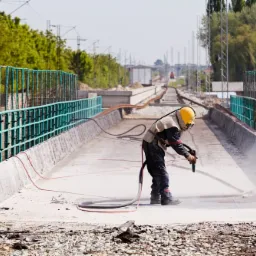 Pracownik w kombinezonie ochronnym i żółtym hełmie czyści betonową powierzchnię mostu za pomocą strumienia ściernego, widoczne zabezpieczenia mostu i tory kolejowe w tle.