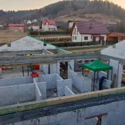 Budowa domu z białych bloczków, widoczne ściany w stanie surowym otwartym, pracownik pod zielonym namiotem tnie bloczki, w tle zabudowa jednorodzinna i zalesione wzgórza.