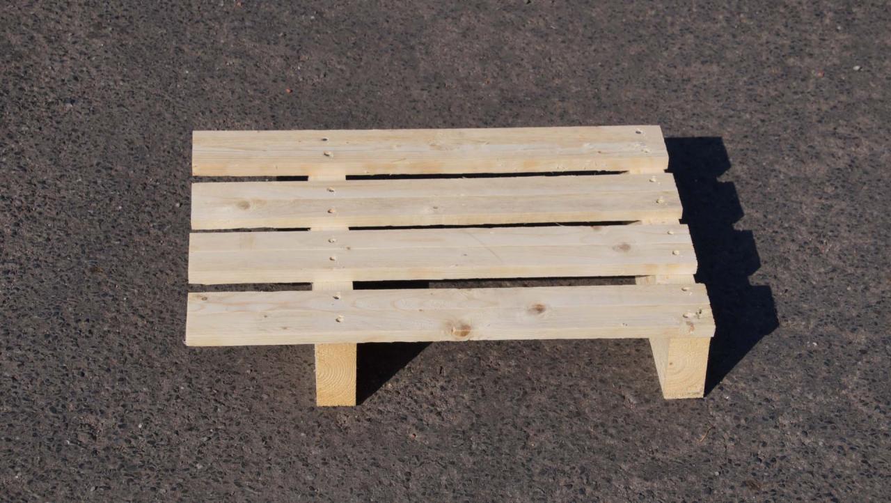 Jasna, surowa paleta z drewna na szorstkim, ciemnym tle. Proste deski połączone w regularny wzór, wsparte na dwóch kantówkach. Widoczne słoje i naturalne niedoskonałości drewna.