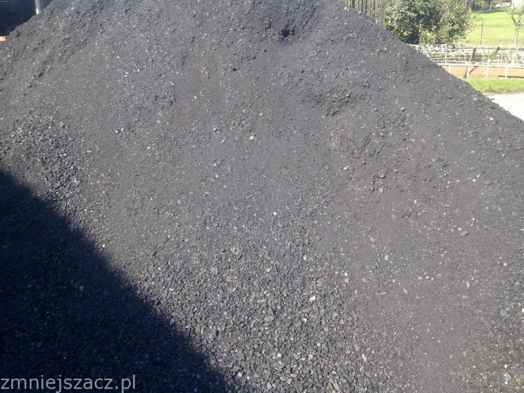 Góra czarnego miału węglowego o nieregularnej powierzchni, z widocznym cieniem po lewej stronie i fragmentem ogrodzenia w tle.