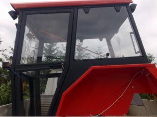 Część kabiny ciągnika rolniczego z widocznymi szybami, czarną ramą i czerwonymi elementami karoserii, na tle zieleni i pochmurnego nieba.